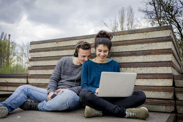 Glückliches Teenager-Paar, das mit Laptop und Kopfhörern im Freien sitzt - EGBF000137