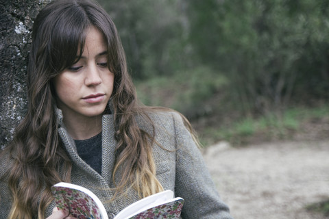 Junge Frau liest ein Buch und lehnt sich an eine Korkeiche, lizenzfreies Stockfoto