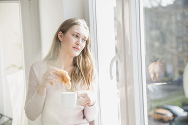 Portrait of young woman with croissant and cup of coffee looking through window - FMKF002447