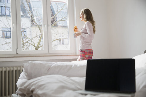 Junge Frau mit Croissant und Tasse Kaffee steht im Schlafzimmer und schaut durch das Fenster - FMKF002448