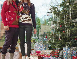Couple standing in front of Christmas tree wearing Christmas jumpers - RHF001326