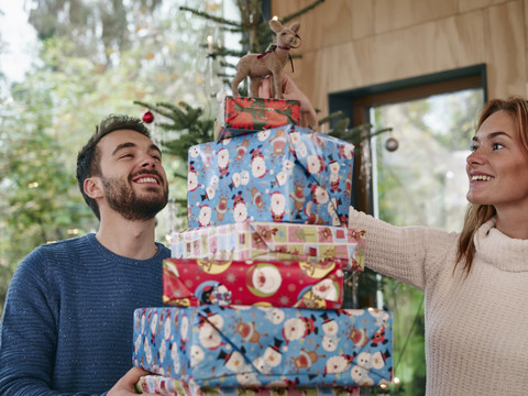 mann trägt einen Stapel Weihnachtsgeschenke, Frau legt ein Rentier oben drauf, lizenzfreies Stockfoto