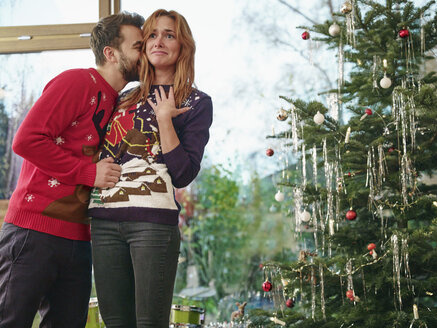 Paar steht vor Weihnachtsbaum, Mann küsst überraschte Frau - RHF001329