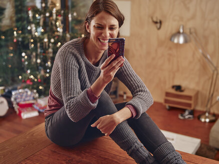 Junge Frau sitzt auf dem Boden vor einem Weihnachtsbaum und benutzt ein Smartphone - RHF001333