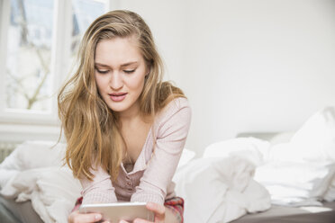 Junge Frau sitzt auf ihrem Bett und schaut auf ihr Smartphone - FMKF002450