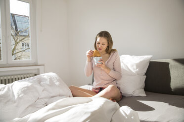 Junge Frau sitzt mit einer Tasse Kaffee und einem Croissant auf ihrem Bett - FMKF002456