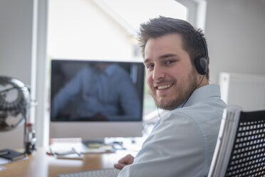 Portrait of smiling young man at desk in office wearing headphones - PAF001613