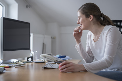 Lächelnde junge Frau am Schreibtisch im Büro, die auf den Computerbildschirm schaut, lizenzfreies Stockfoto