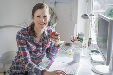 Porträt einer lächelnden jungen Frau am Schreibtisch im Büro, die einen Apfel hält - PAF001619