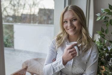 Porträt einer lächelnden jungen Frau, die eine Tasse Kaffee am Fenster hält - PAF001626