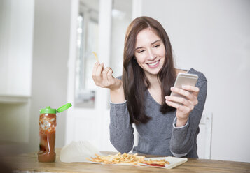 Porträt einer lächelnden jungen Frau, die ein Smartphone in der Hand hält und Pommes frites mit Ketchup isst - FMKF002477