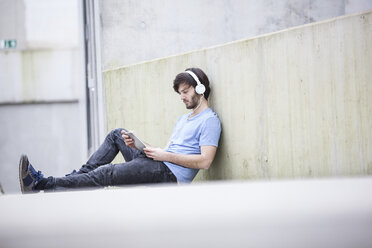 Young man sitting on the ground listening music with headphones looking at digital tablet - FMKF002488