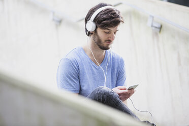 Junger Mann hört Musik mit Kopfhörern und schaut auf sein Smartphone - FMKF002496