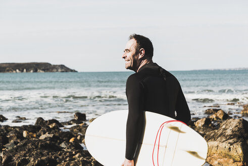 Frankreich, Bretagne, Finistere, Halbinsel Crozon, lächelnder Mann am felsigen Strand mit Surfbrett - UUF006731