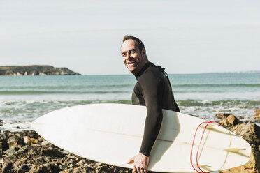 Frankreich, Bretagne, Finistere, Halbinsel Crozon, glücklicher Mann am felsigen Strand mit Surfbrett - UUF006732