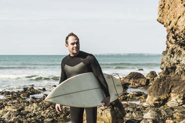 Frankreich, Bretagne, Finistere, Halbinsel Crozon, selbstbewusster Mann am felsigen Strand mit Surfbrett - UUF006733