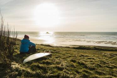 Frankreich, Bretagne, Finistere, Halbinsel Crozon, Frau an der Küste sitzend bei Sonnenuntergang mit Surfbrett - UUF006744