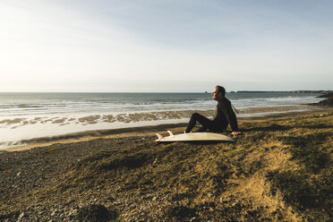 Frankreich, Bretagne, Finistere, Halbinsel Crozon, Mann sitzt mit Surfbrett an der Küste - UUF006749