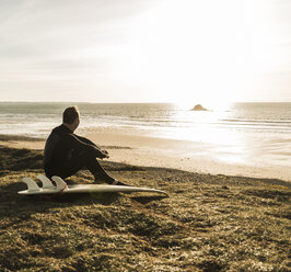 Frankreich, Bretagne, Finistere, Halbinsel Crozon, Mann sitzt bei Sonnenuntergang mit Surfbrett an der Küste - UUF006750