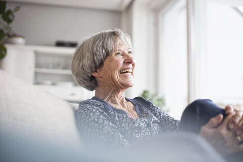 Porträt einer lachenden älteren Frau, die zu Hause auf der Couch sitzt - RBF004107