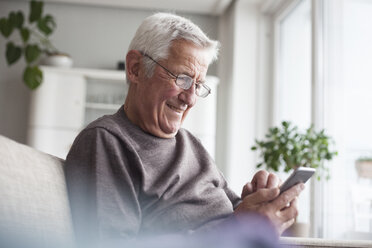 Portrait of smiling senior man sitting on couch at home using smartphone - RBF004118