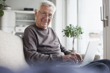 Portrait of smiling senior man sitting on couch at home using laptop - RBF004121