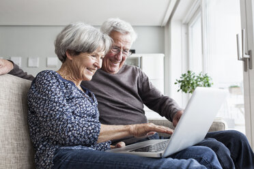 Glückliches älteres Paar sitzt auf der Couch im Wohnzimmer und benutzt einen Laptop - RBF004122