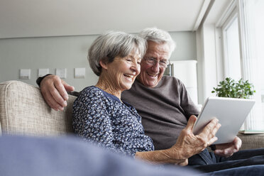 Glückliches älteres Paar, das auf der Couch im Wohnzimmer sitzt und ein digitales Tablet benutzt - RBF004123