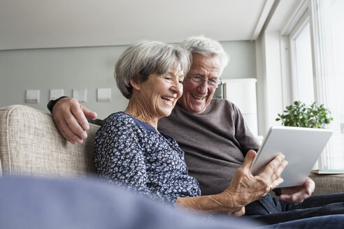Happy senior couple sitting on the couch in the living room using digital tablet - RBF004123