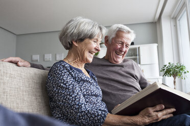 Lachendes älteres Paar, das auf der Couch im Wohnzimmer sitzt und sich ein Fotoalbum ansieht - RBF004124