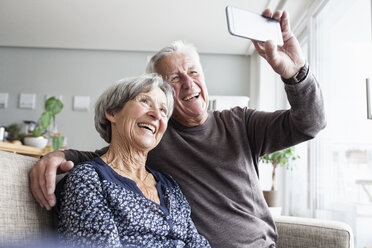 Laughing senior couple sitting on the couch in the living room taking selfie with smartphone - RBF004128