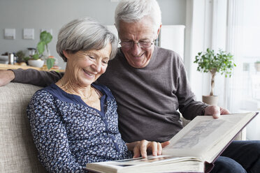 Happy senior couple sitting on the couch of living room watching photo album - RBF004129