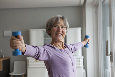 Portrait of happy senior woman doing fitness exercise with dumbbells at home - RBF004153