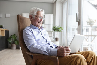 Portrait of senior man sitting on armchair at home using laptop - RBF004158