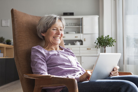 Porträt einer älteren Frau, die zu Hause auf einem Sessel sitzt und ein digitales Tablet benutzt, lizenzfreies Stockfoto