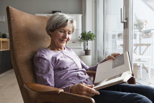 Portrait of senior woman sitting on armchair at home watching photo album - RBF004167