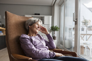 Portrait of senior woman sitting on armchair at home looking through window - RBF004168