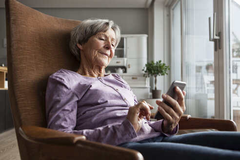 Porträt einer älteren Frau, die zu Hause auf einem Sessel sitzt und ein Smartphone benutzt - RBF004170
