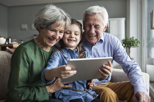 Grandparents and their granddaughter sitting together on the couch looking at digital tablet - RBF004204