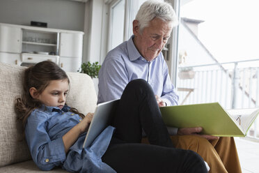 Großvater und seine Enkelin sitzen zusammen auf der Couch mit Buch und digitalem Tablet - RBF004209