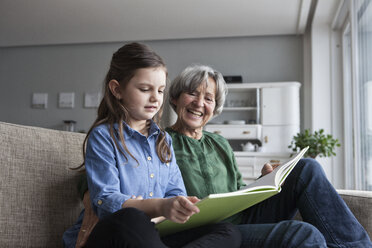 Grandmother and her granddaughter sitting together on the couch with a book - RBF004212