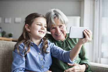 Grandmother and her granddaughter sitting together on the couch taking selfie with smartphone - RBF004221