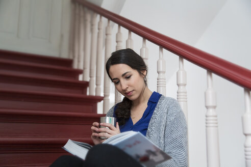 Frau im Treppenhaus, die eine Tasse Kaffee trinkt und ein Buch liest - FKF001687