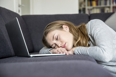 Junge Frau mit Laptop, die sich zu Hause auf ihrer Couch entspannt - FMKF002498