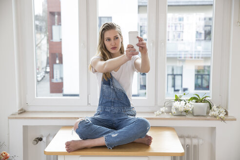 Junge Frau sitzt auf einem Tisch zu Hause und macht ein Selfie mit ihrem Smartphone - FMKF002499