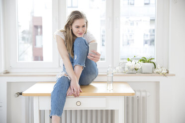 Young woman sitting on a table at home looking at smartphone - FMKF002501