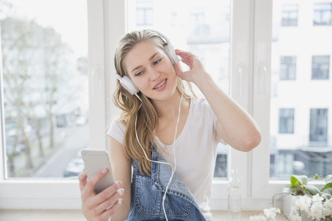 Porträt einer jungen Frau, die mit Kopfhörern vor einem Fenster Musik hört, lizenzfreies Stockfoto