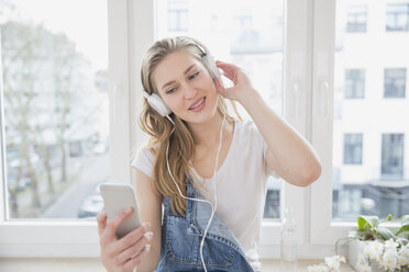 Porträt einer jungen Frau, die mit Kopfhörern vor einem Fenster Musik hört - FMKF002502
