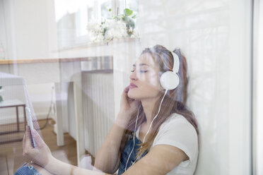 Junge Frau sitzt hinter einer Fensterscheibe und hört mit Kopfhörern Musik - FMKF002503