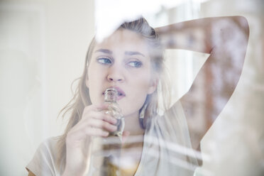 Porträt einer jungen Frau, die durch eine Fensterscheibe schaut und Wasser trinkt - FMKF002505